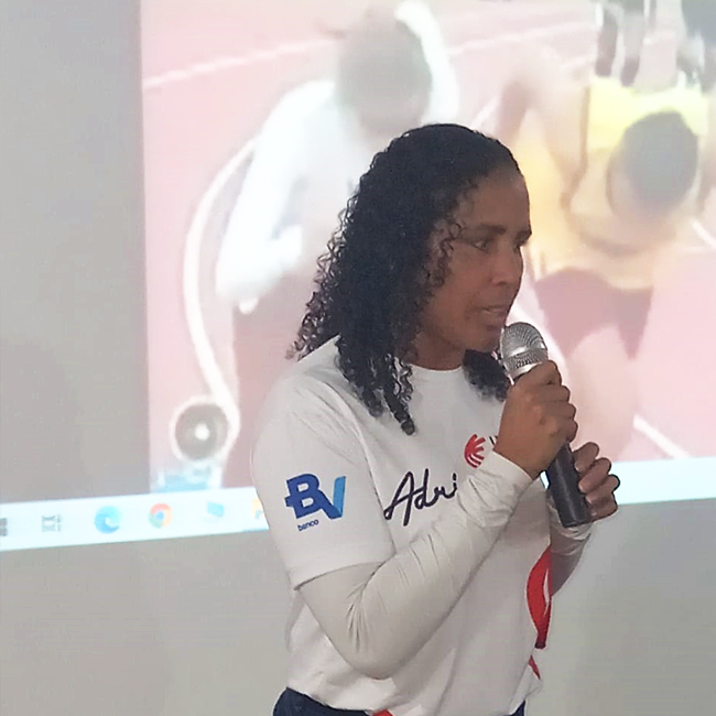 Adria fala ao microfone em frente à um telão que mostra foto de atletismo.