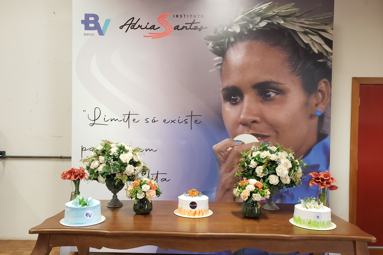 Mesa com bolos e flores. Ao fundo painel com foto da Adria e logotipos do Banco BV e do Instituto.