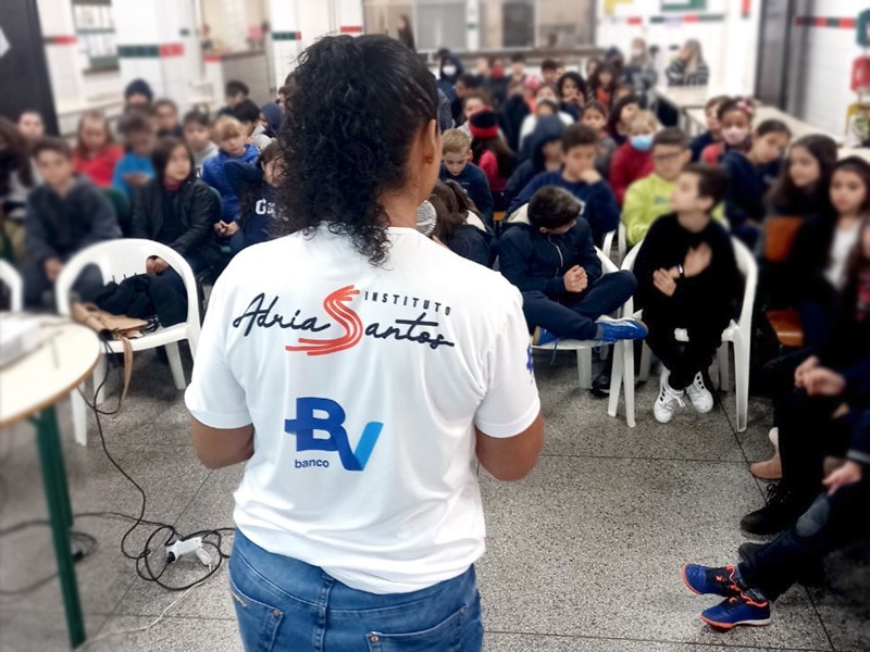 Adria veste camiseta do Instituto e fala diante de sala cheia de crianças.