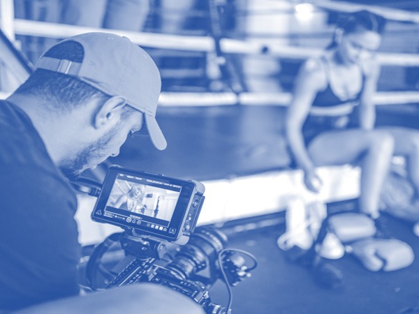 Photo in blue tone of a man filming a woman and looking at the image through the screen of a camera.