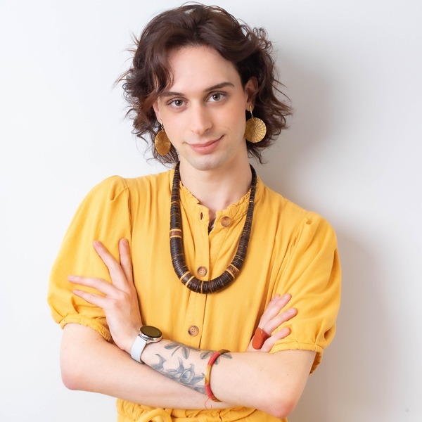 Photo of Vinicius, white skin, short hair, wears a yellow shirt, necklace and has his arms crossed