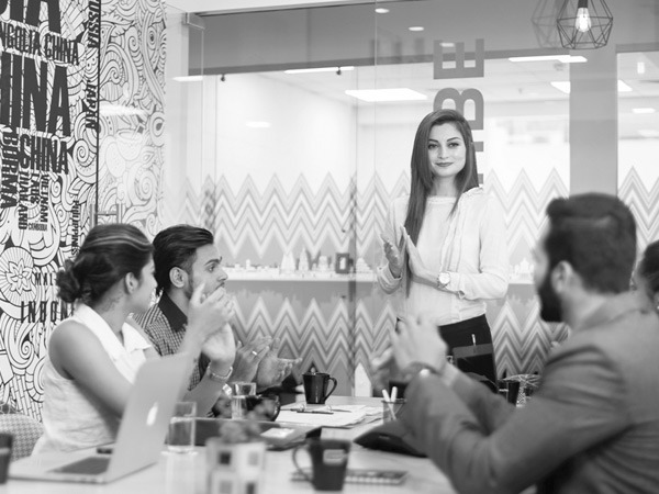 Black and white photo of people in a meeting. A woman is standing with her hands together.