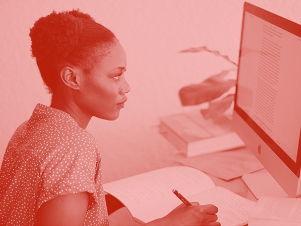 Foto em tom vermelho de uma mulher negra, com blusa de bolinhas, trabalhando em um computador.
