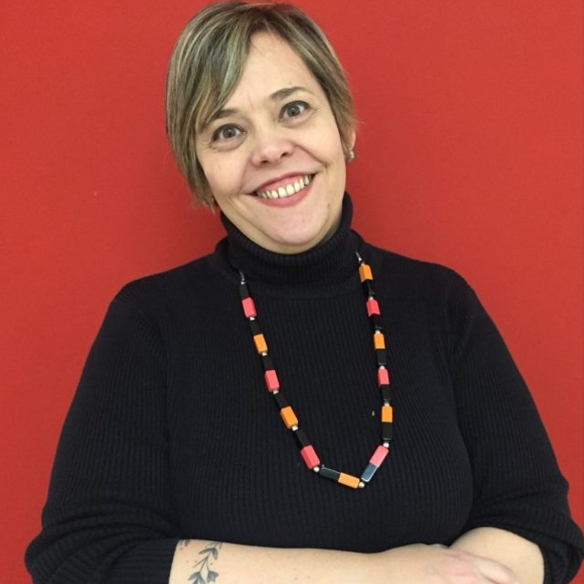 White woman, straight and short hair. She wears a black woolen blouse and a necklace.