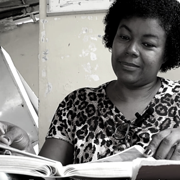 Foto branco e preta: Mulher negra lê um livro em sala com parece descascada ao fundo.