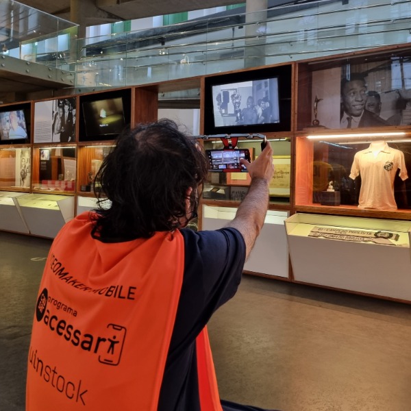 Jovem segurando um celular na mão com um estabilizador, filmando um painel de obra em museu